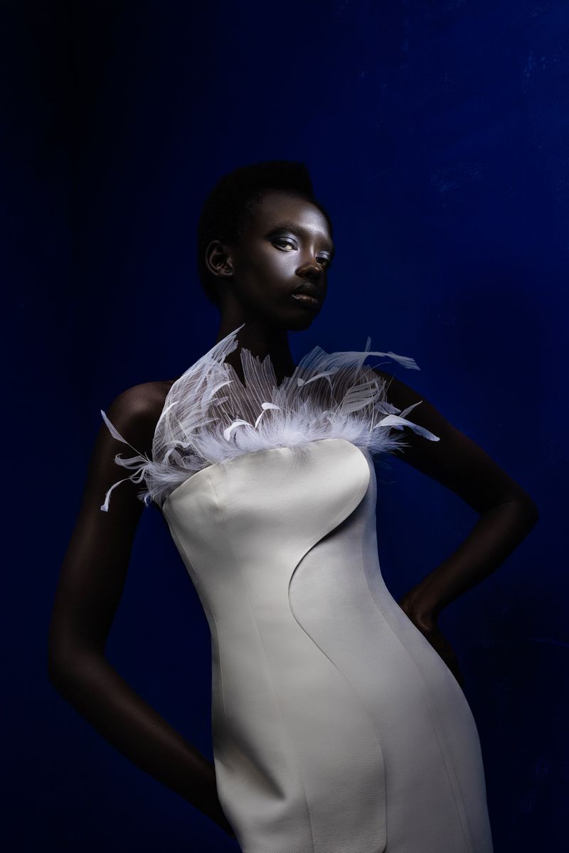 Woman in a pale dress with feather collar poses against a dark blue background in a studio shot emphasizing fashion and beauty.