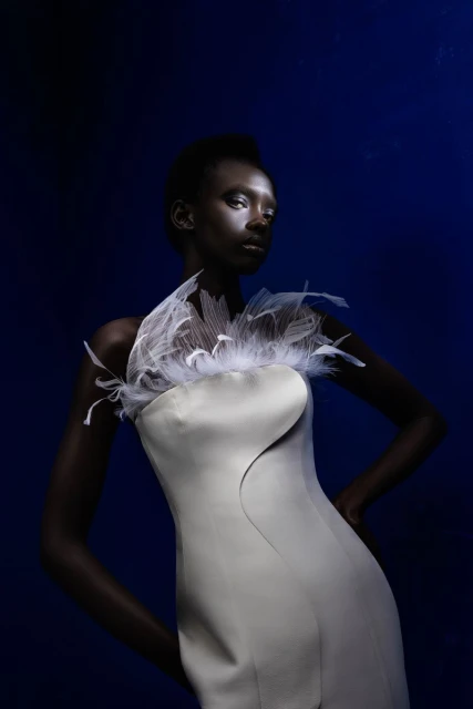 Woman in a pale dress with feather collar poses against a dark blue background in a studio shot emphasizing fashion and beauty.