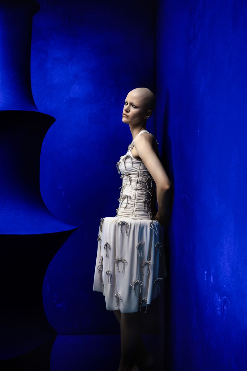 Young woman in stylish short dress poses against a cobalt blue architectural backdrop with unique urn shapes. Fashion editorial.