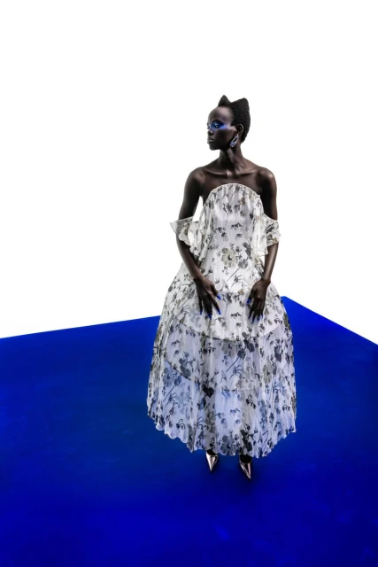 Fashion model wearing a white floral gown with blue eyeshadow and dark lipstick, standing on a blue platform in a studio setting. 