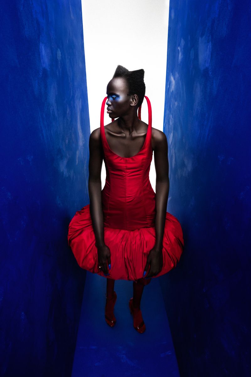 Fashion model in a vibrant red dress with blue eye makeup poses in a narrow corridor with a bright light at the end.