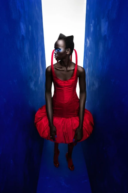 Fashion model in a vibrant red dress with blue eye makeup poses in a narrow corridor with a bright light at the end.