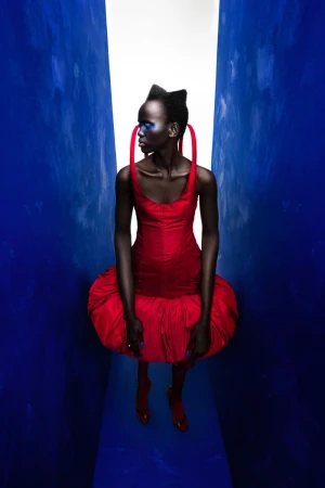 Fashion model in a vibrant red dress with blue eye makeup poses in a narrow corridor with a bright light at the end.