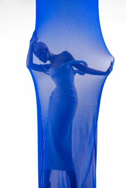 Elegant, bald woman in a white dress poses in a photo studio, framed by sheer royal blue fabric. Fashionable, minimalist composition.