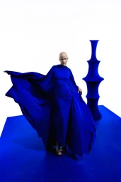 Model in vibrant blue dress with flowing cape standing on a blue platform against a white background