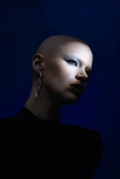Close-up portrait of a bald model wearing silver earrings against a dark blue background for Numero Netherlands INTO THE BLUE editorial.