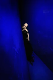 Model Coco Labbee with a shaved head wearing a black gown leaning between deep blue angled walls.