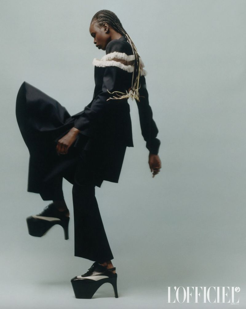 Fashion-forward woman strikes a pose, showcasing a stylish black suit and platform heels against a muted light blue backdrop in studio.