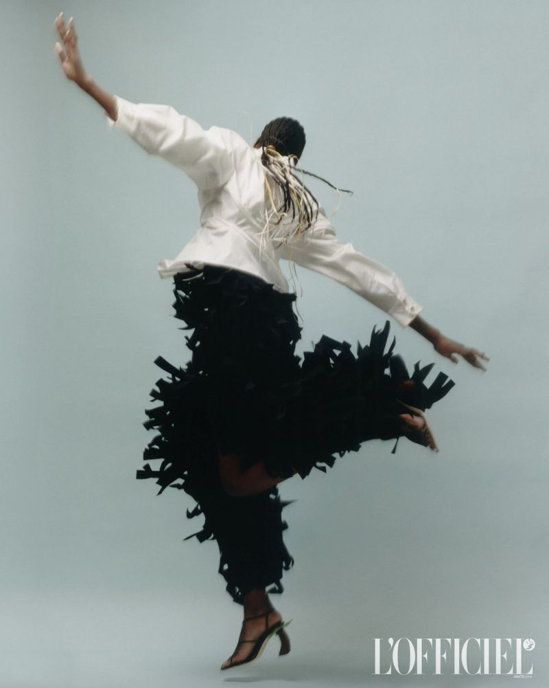A woman in a white top and fringe skirt leaps in a studio setting, embodying fashion and style, as captured by L'Officiel magazine.