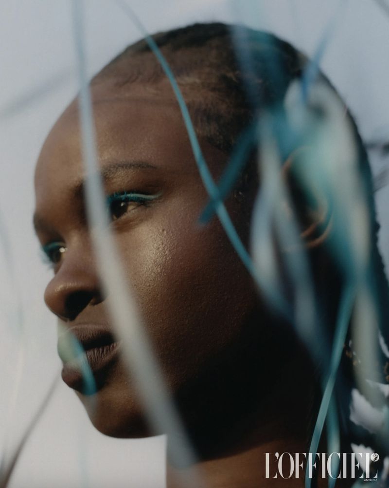 Close-up shot of a Black woman with blue eyeliner. Blue decorative objects are in the foreground and the L'Officiel magazine logo is visible.