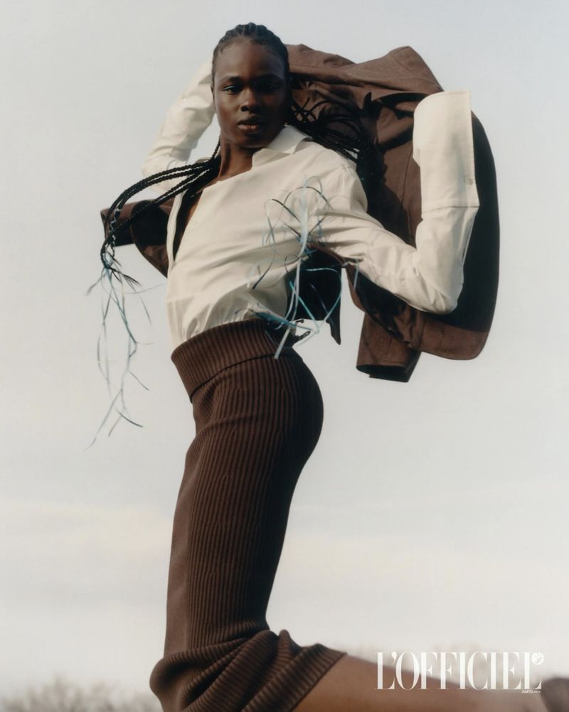 Fashion editorial shot of an adult female model wearing a white long-sleeved top, brown skirt, and jacket against a light sky background.