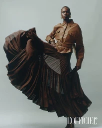 Model Bakhita Lual wearing a brown belted jacket and flowing tiered skirt against a seamless grey backdrop for L'Officiel Italia