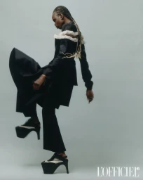 Model in black platform shoes and dark outfit lifting leg against grey seamless backdrop for L'Officiel Italia.