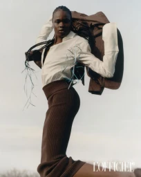 Model wearing a white shirt and brown skirt holding a brown jacket against a pale sky background for L'Officiel Italia
