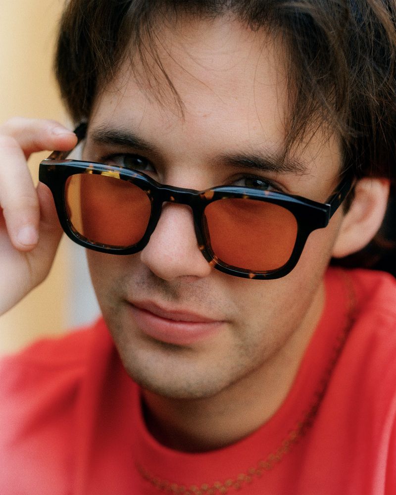 Close-up portrait of a young adult male wearing tortoise-shell glasses with orange lenses, gazing directly at the viewer in an indoor setting.