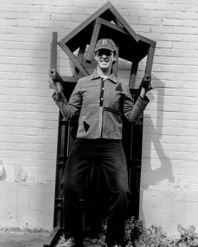 Monochrome shot of an adult wearing a baseball cap, jacket, and jeans sitting in a metal frame against a brick wall outdoors.