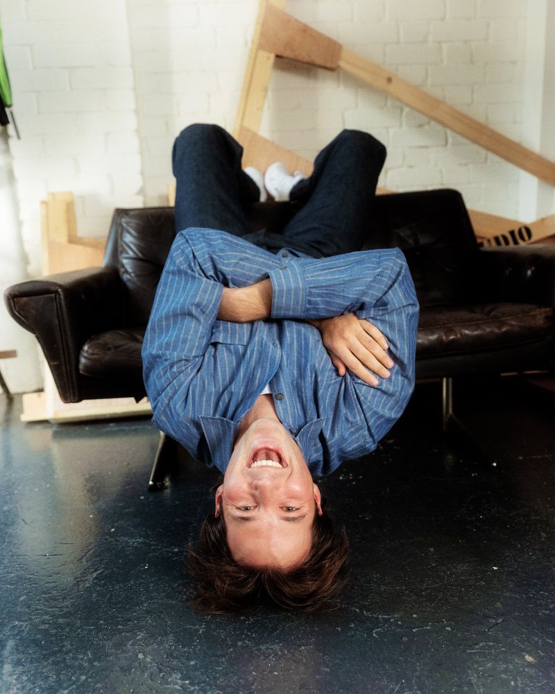 A person is upside down on a brown leather couch with white wall and black floors inside. They are wearing blue jeans.