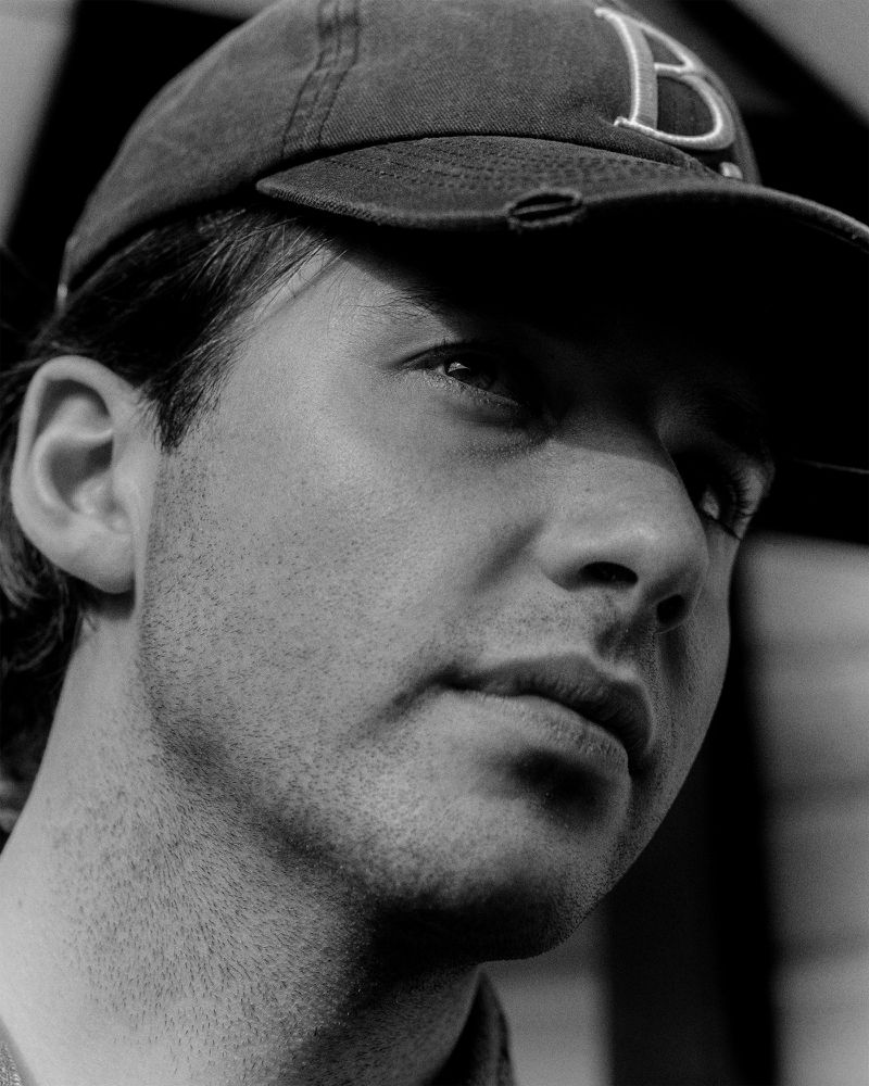 Black and white portrait of a man wearing a baseball cap looking off-camera with neutral expression, close-up studio shot.