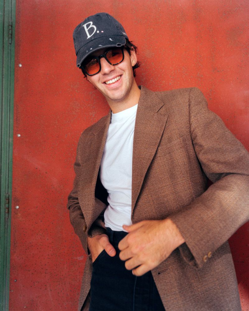 Young man in a brown blazer, white t-shirt, and baseball cap smiles in front of a red-painted metal wall with green trim in daylight.