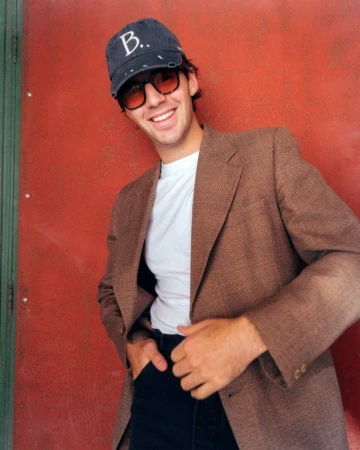 Young man in a brown blazer, white t-shirt, and baseball cap smiles in front of a red-painted metal wall with green trim in daylight.