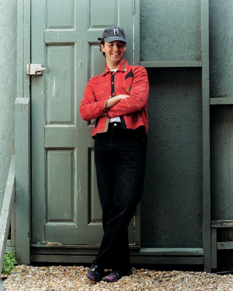 Full shot of an adult man wearing a baseball cap, red jacket, and black pants standing outdoors near a door and stone ground.
