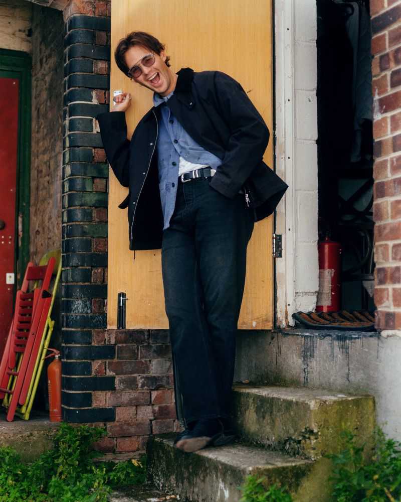 Smiling man with sunglasses leans against a yellow door on a brick porch next to a red folding chair and fire extinguisher.