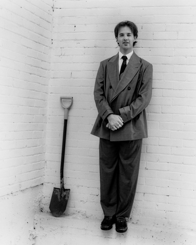 Adult male in double-breasted suit poses next to a shovel, leaning against a white brick wall in an indoor setting.  Black and white image.