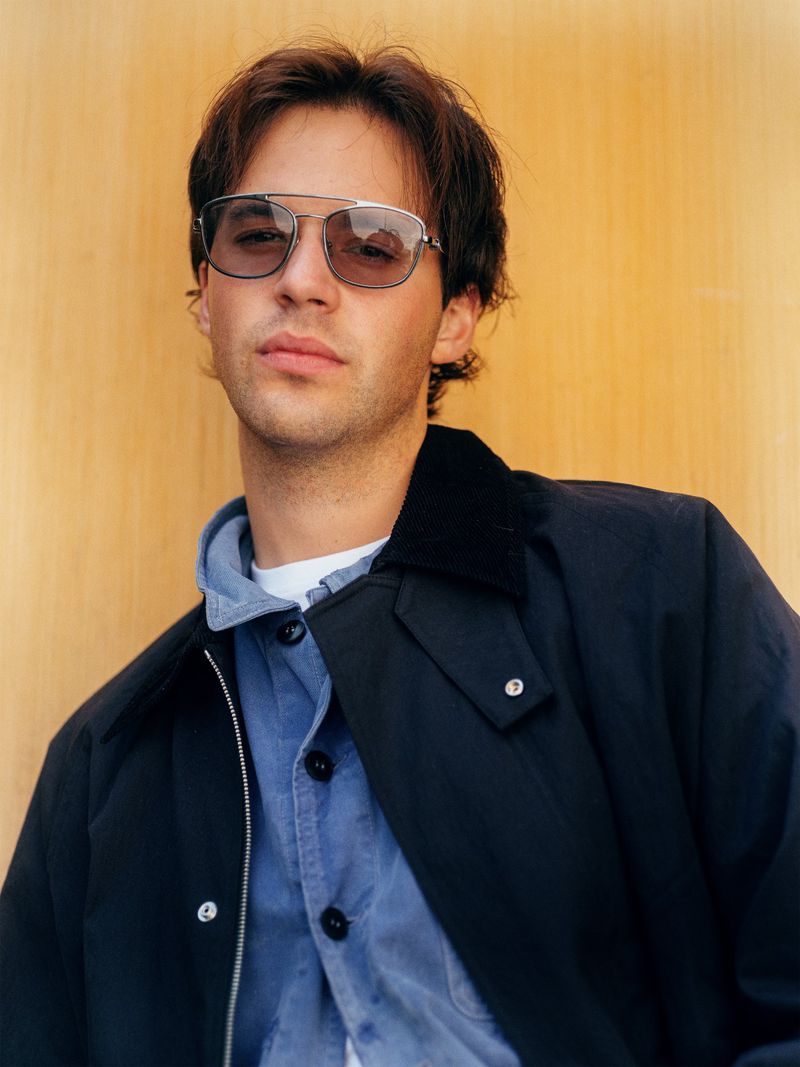 Portrait of a light-skinned adult male wearing sunglasses, a blue shirt and a dark jacket, posing against a light wood background.