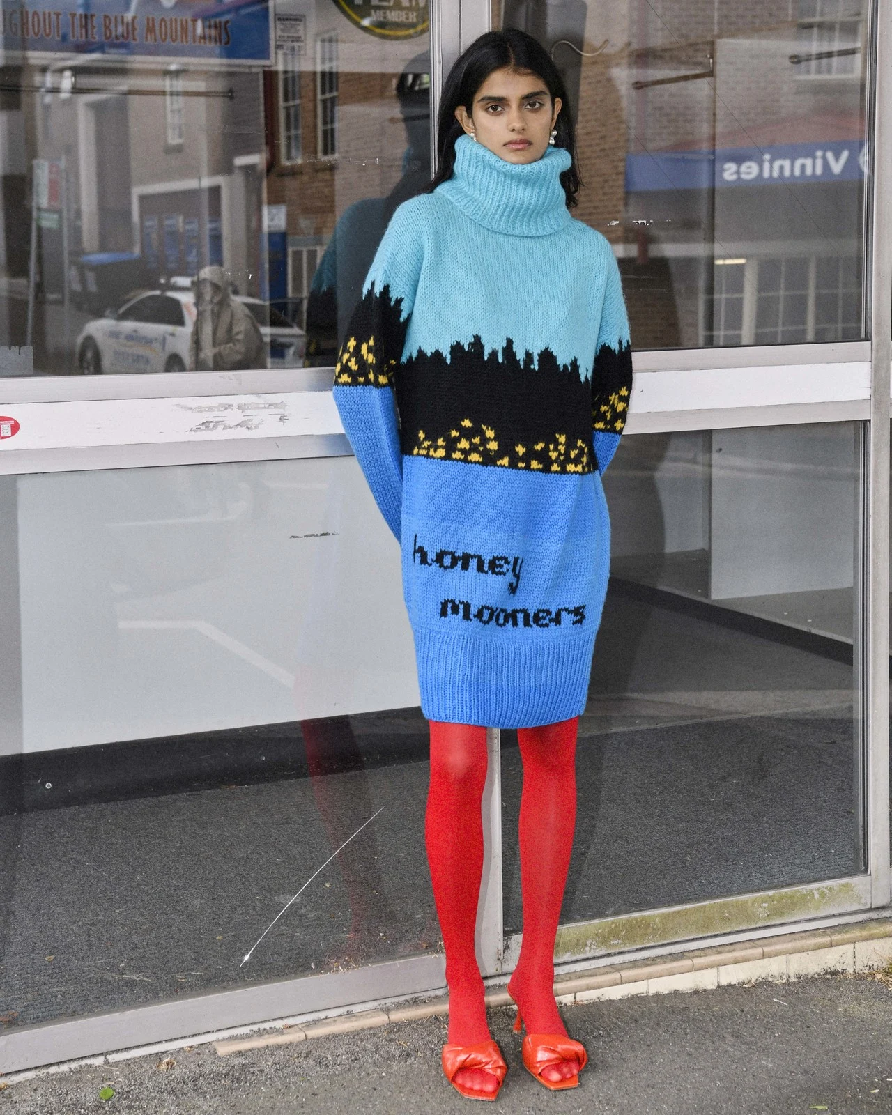 Woman wearing a blue turtleneck dress with red tights and orange shoes is standing in front of a building outdoors.