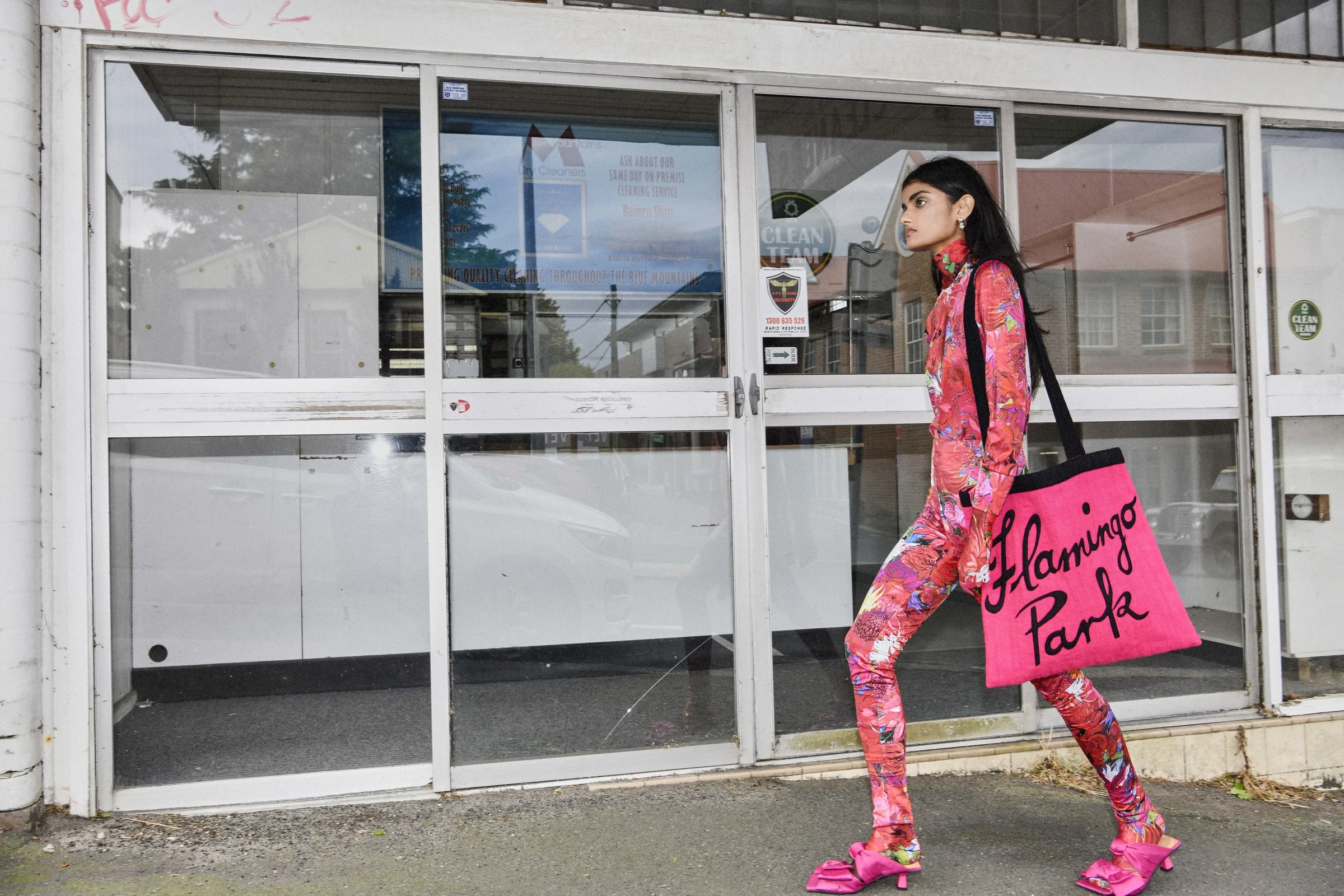 A woman in floral pantsuit walking outdoors carrying a pink tote bag. She is walking past a row of commercial doors with glass panes.
