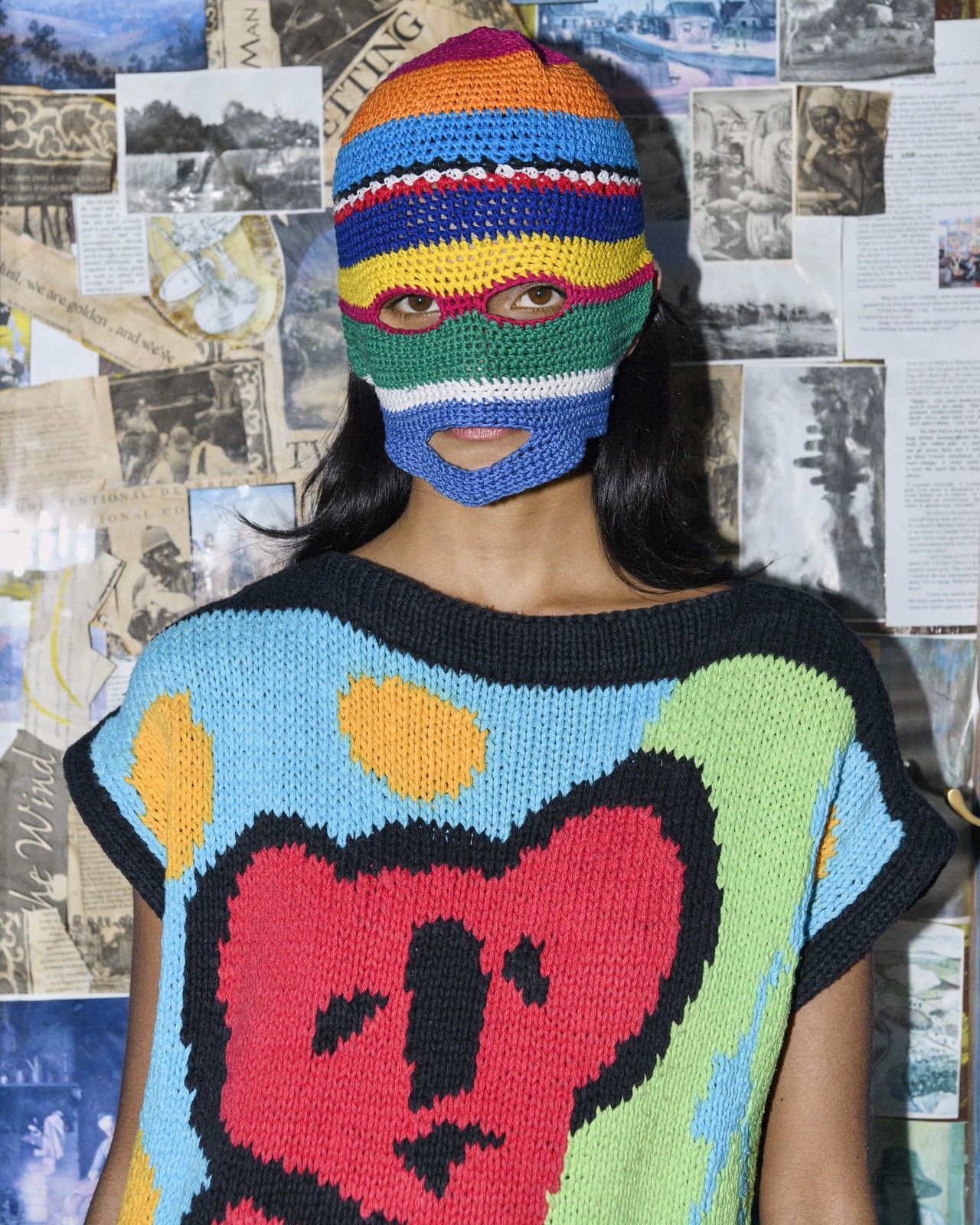 A person wearing a colorful knit balaclava and a colorful knit sweater with a bear design, against a background of vintage newspaper clippings.