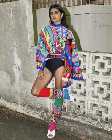 An adult female poses outdoors wearing a colorful patterned sweater, black bottoms, mismatched leg warmers, and sandals on a concrete path.