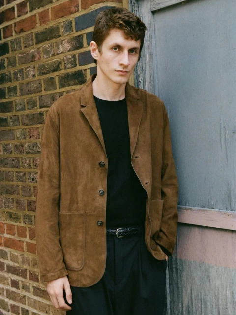 Man in brown suede blazer with black shirt and pants stands near a brick wall and a closed wood door for a fashion editorial.