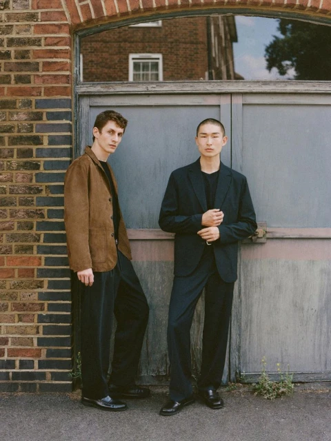Two men pose in fashionable jackets and pants next to a textured brick and metal door in an urban environment during daylight.