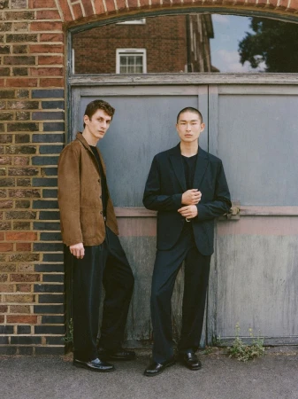 Two men pose in fashionable jackets and pants next to a textured brick and metal door in an urban environment during daylight.