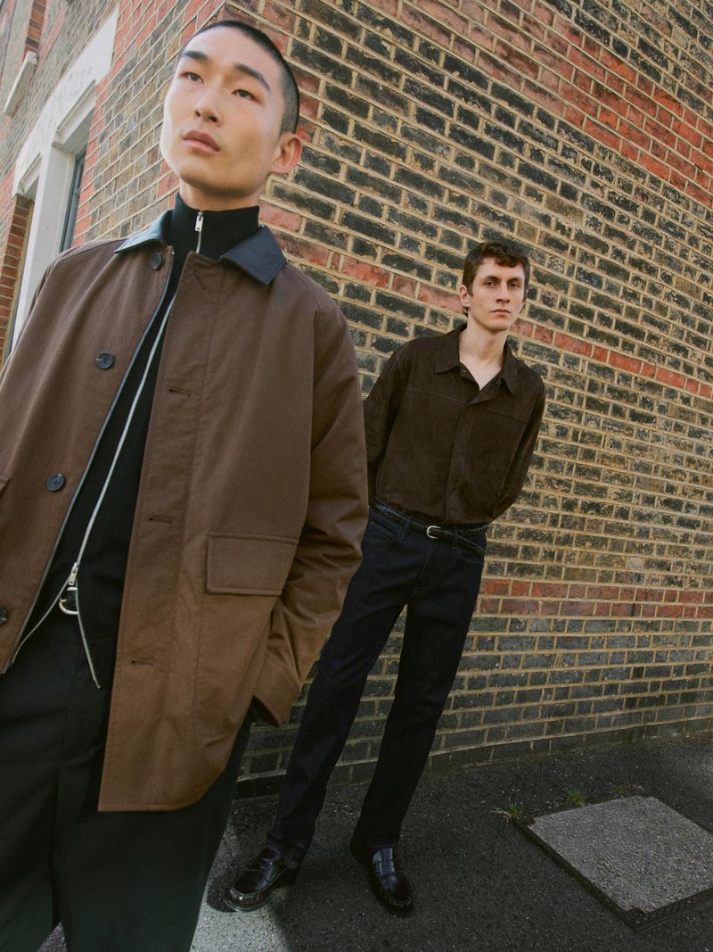 Two young adult men pose outdoors in brown and black attire against a brick wall in daytime, fashion editorial, medium angle.