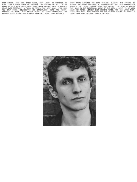 Monochrome portrait of a young adult man with short hair, gazing directly at the camera, set against a blurred brick wall background.