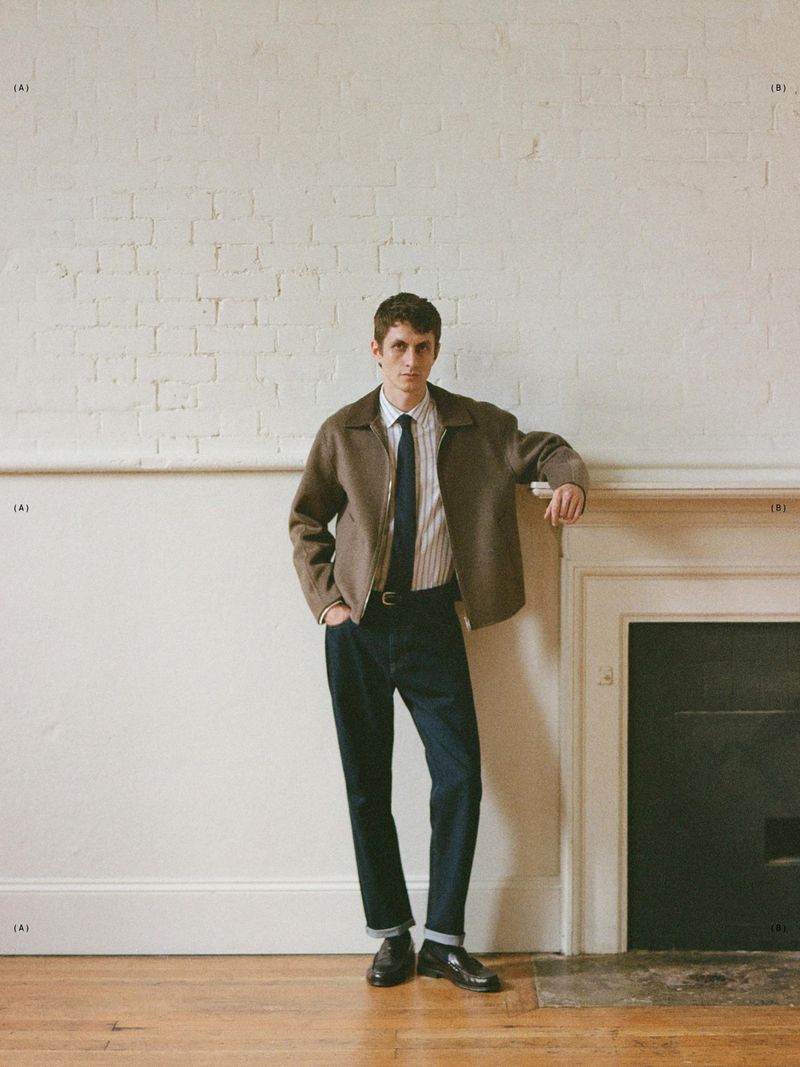 Man in brown jacket and tie leans against a fireplace. Indoor fashion portrait on wood floor in front of white brick wall.
