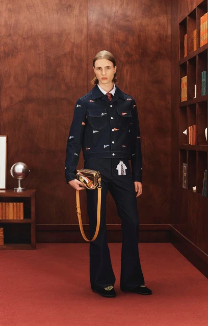 A fashion shot of a slender, fair-skinned young adult in a blue patterned denim suit, holding a handbag in an indoor library setting.