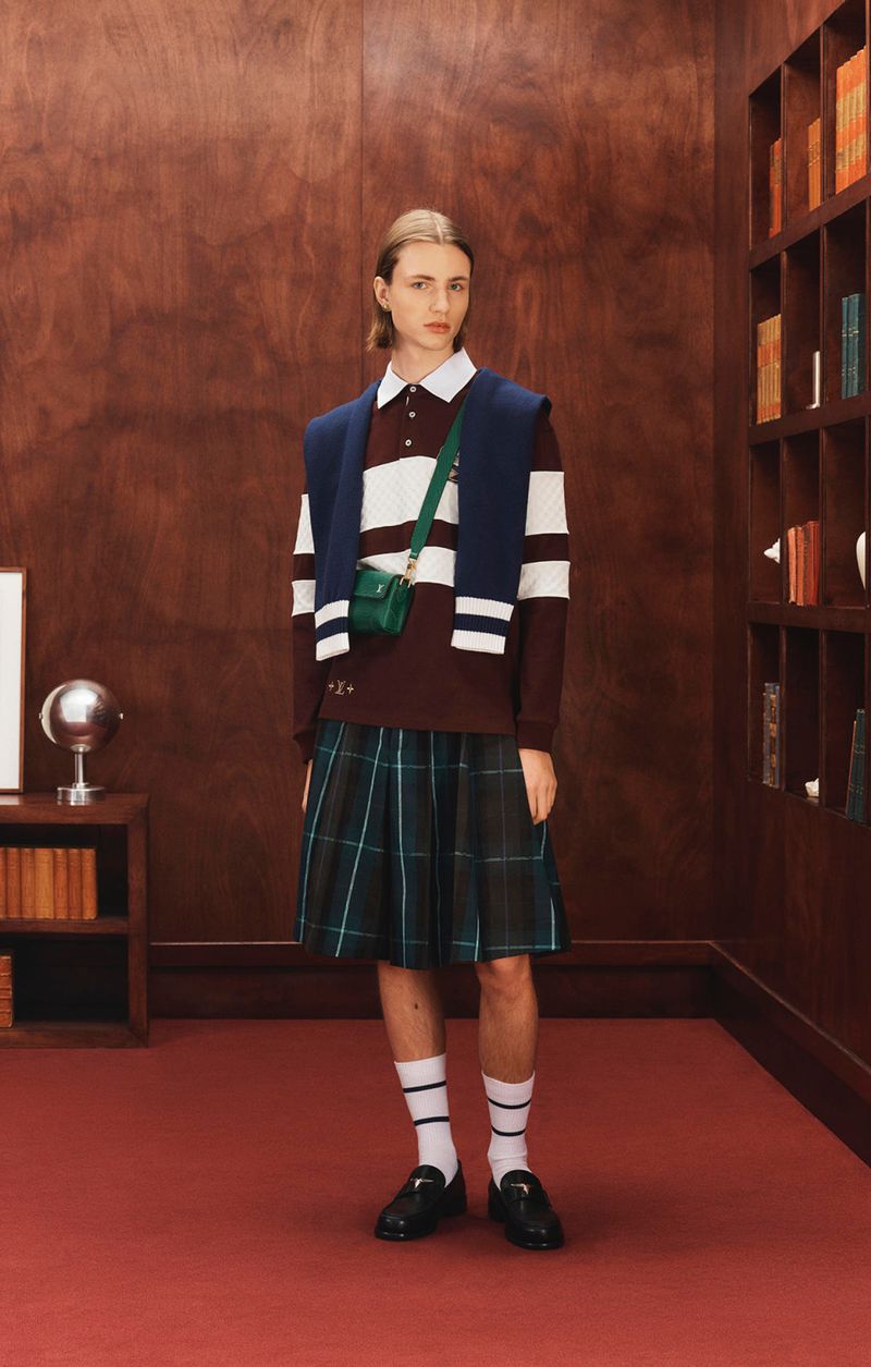 Full-length shot of a slender young person wearing a plaid skirt and striped rugby shirt standing in a room with a bookcase.