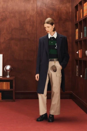Male fashion model poses in a library-like setting wearing a navy coat, striped polo, wide-legged khaki pants, and black dress shoes.