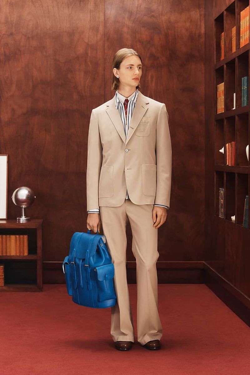 Man in a suit stands in a library setting, holding a blue backpack. The image highlights fashion and classic style.