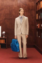 Model standing in a beige suit holding a vibrant blue leather backpack inside a wood-paneled room