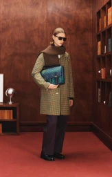 Model Indiana Van't Slot wearing a check coat, brown knit scarf, sunglasses, and a monogram crossbody bag in a wood-paneled room.