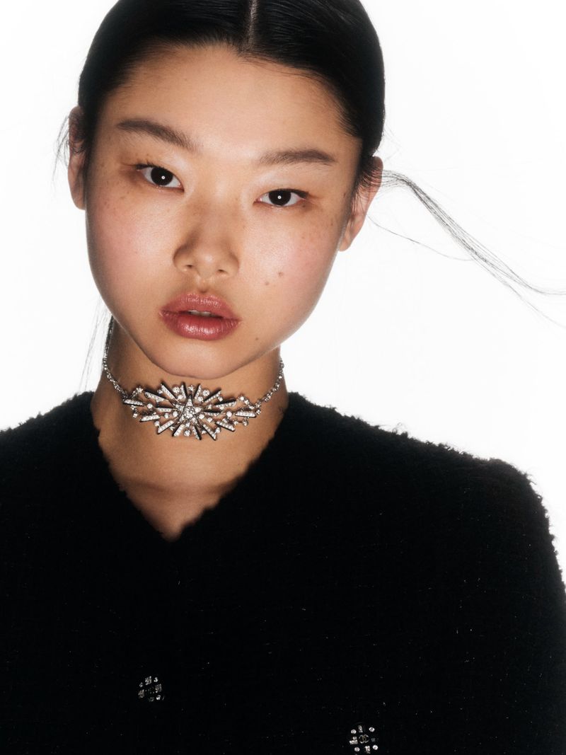 Close-up fashion shot of a young Asian woman wearing an ornamental choker necklace and a black tweed jacket against a white backdrop.