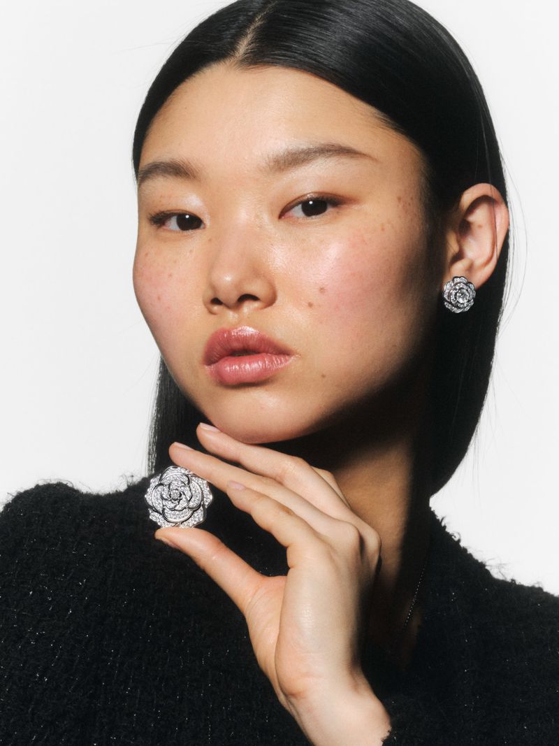 Close-up of an Asian woman wearing rose earrings and a black tweed jacket holding a rose pin in a bright, neutral studio shot.