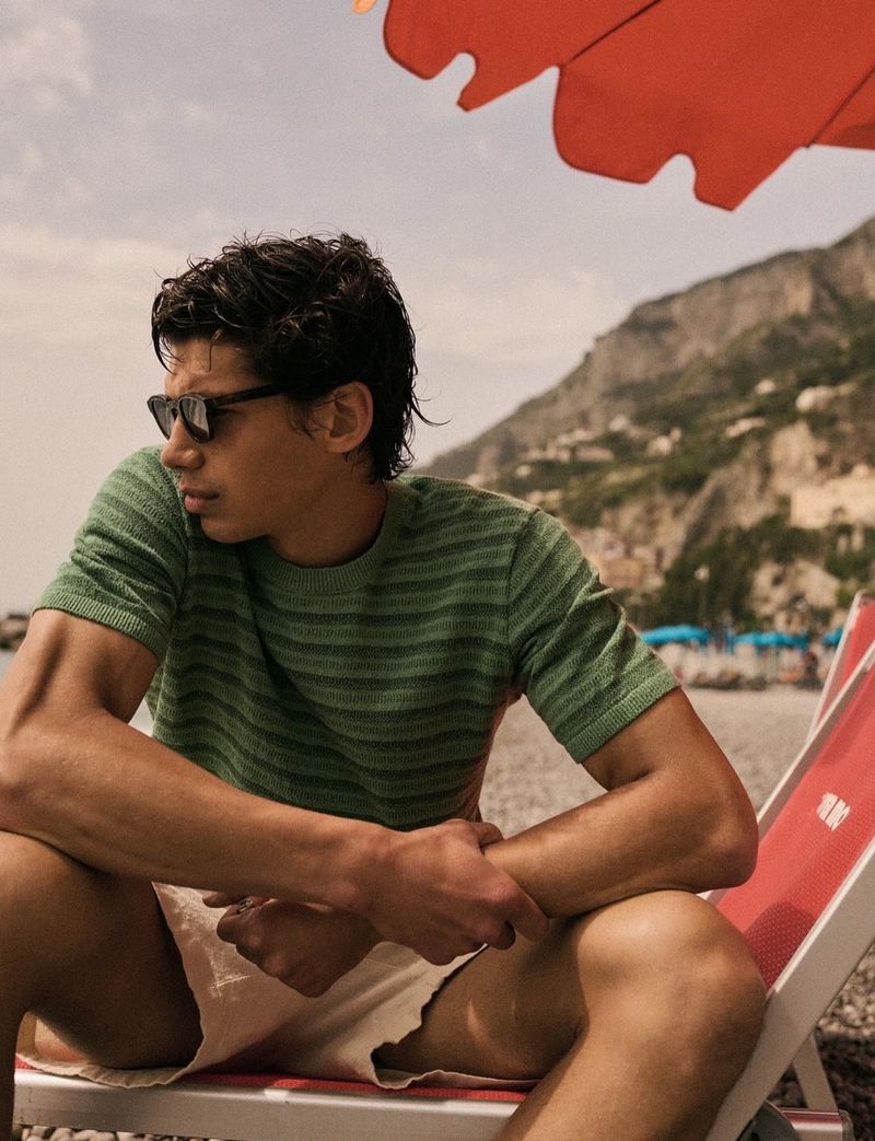 Man in a green short-sleeve knit shirt and sunglasses sitting on a red beach chair beneath an orange umbrella.