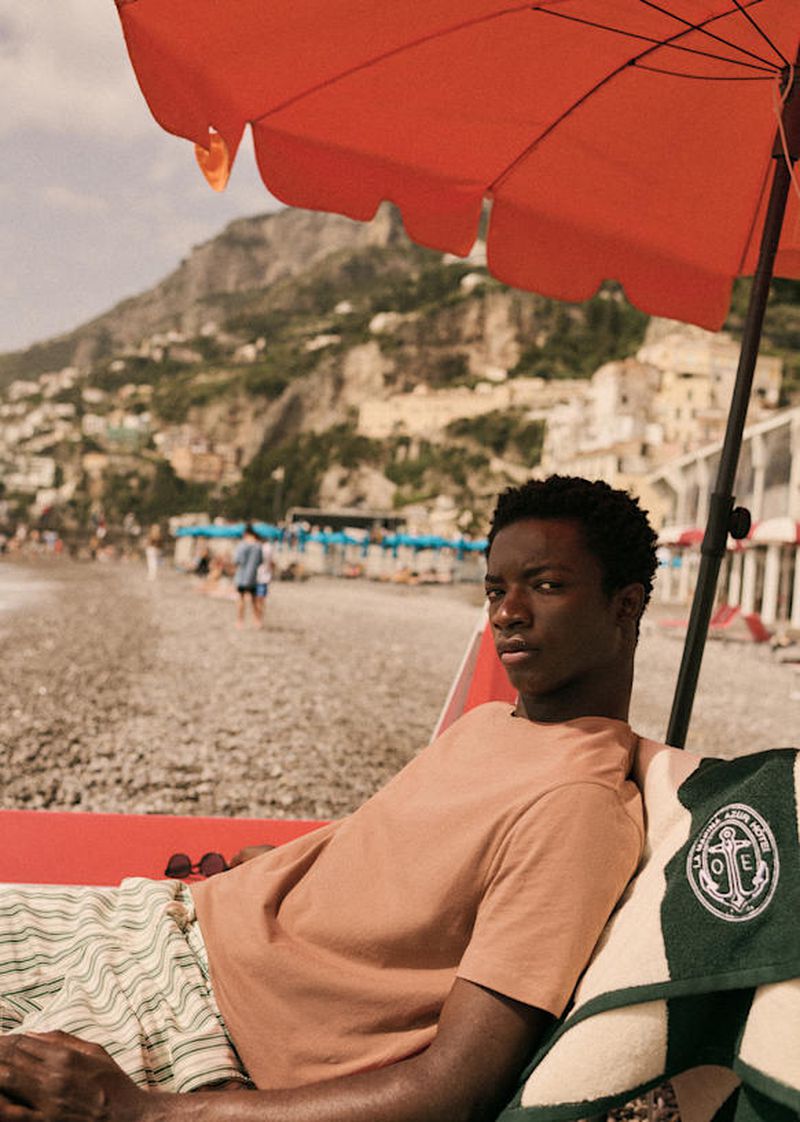 A young man relaxing in a lounge chair on a beach under an orange umbrella, with a beach, cliffs, and town in the background.