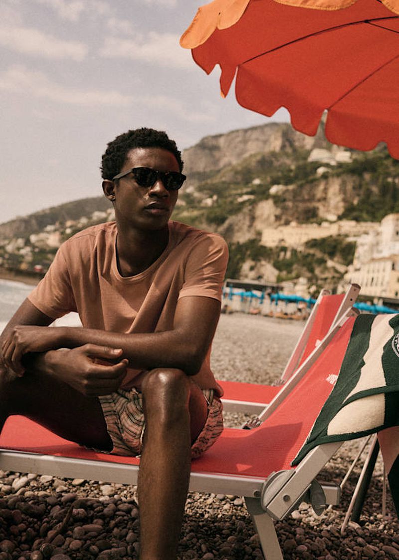 Man in sunglasses and beachwear lounges on a red deck chair on a rocky beach under an orange umbrella with a coastal mountain backdrop.