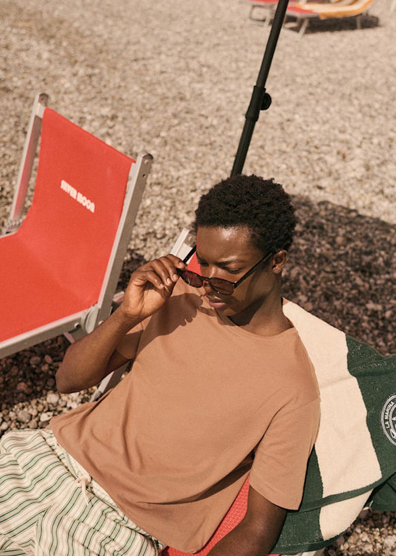Young man in sunglasses lounges on a beach chair near the sea, wearing a t-shirt and striped shorts on a sunny day.